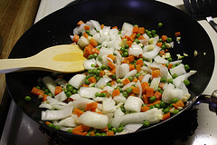 Chopped onions, carrots, and peas being stir fried in a wok with a wooden spatula