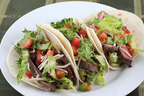 Grilled steak tacos filled with lettuce, tomato, onion, and cheese on a white plate