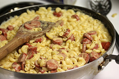 Skillet of macaroni and cheese with sliced hot dogs and wooden spoon