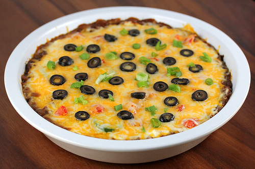 Hot taco dip in a white casserole dish topped with melted Colby‑Jack cheese, sliced tomatoes, olives, onion, and jalapeños