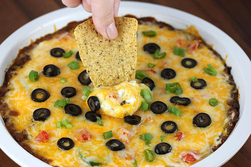 Hot taco dip in a white casserole dish topped with melted Colby‑Jack cheese, sliced tomatoes, olives, onion, and jalapeños