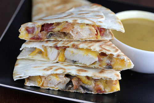 Quesadilla slices filled with chicken, cheese, and bacon on a black plate with dipping sauce