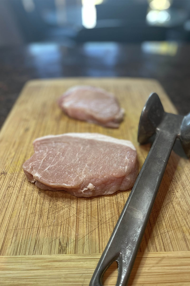 Raw pork loin chops on cutting board with mallet.