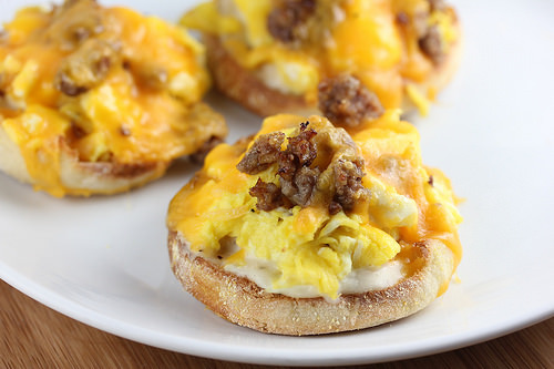 English muffin halves topped with eggs, sausage, and melted cheddar cheese on a white plate