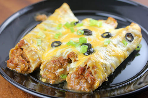 Ground beef enchiladas topped with melted cheese black olives and green onions in baking dish