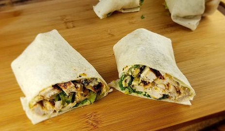 Sliced burrito filled with grilled chicken, lettuce, tomato, and avocado on a wooden board