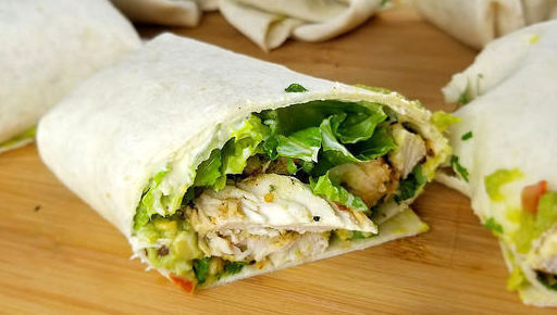 Sliced burrito filled with grilled chicken, lettuce, tomato, and avocado on a wooden board