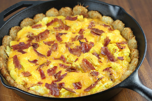 Cast iron skillet with tater tot breakfast pizza topped with scrambled eggs, bacon, and melted cheddar cheese
