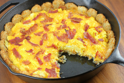 Tater tot breakfast pizza in skillet with a slice removed, revealing eggs, cheese, and bacon filling