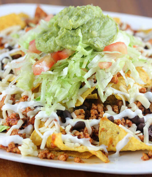 Plate of nachos topped with seasoned chicken, lettuce, tomato, cheese, and guacamole