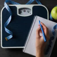 View of hand taking notes on pad of paper near scale for weight loss.