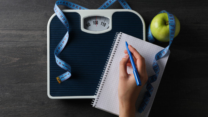 View of hand taking notes on pad of paper near scale for weight loss.