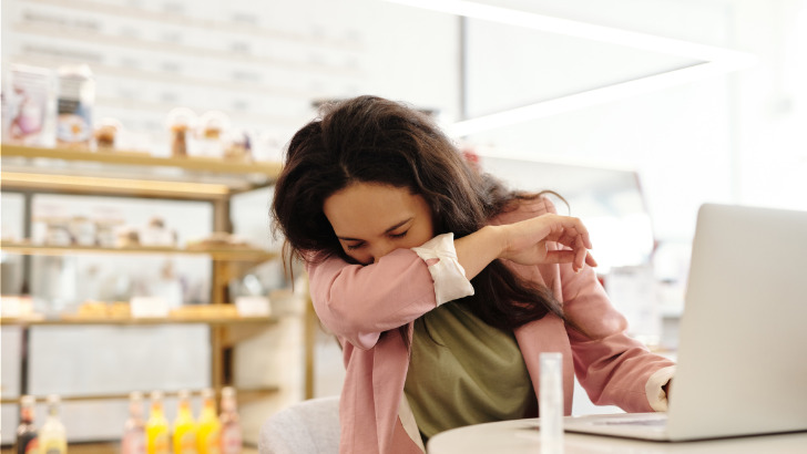 Woman sitting at desk coughs into elbow.