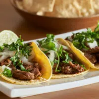 Three carna asada tacos on a serving dish.