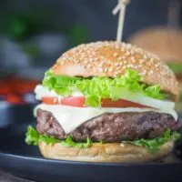 Side view of prepared hamburger on bun with cheese, lettuce, and tomato.