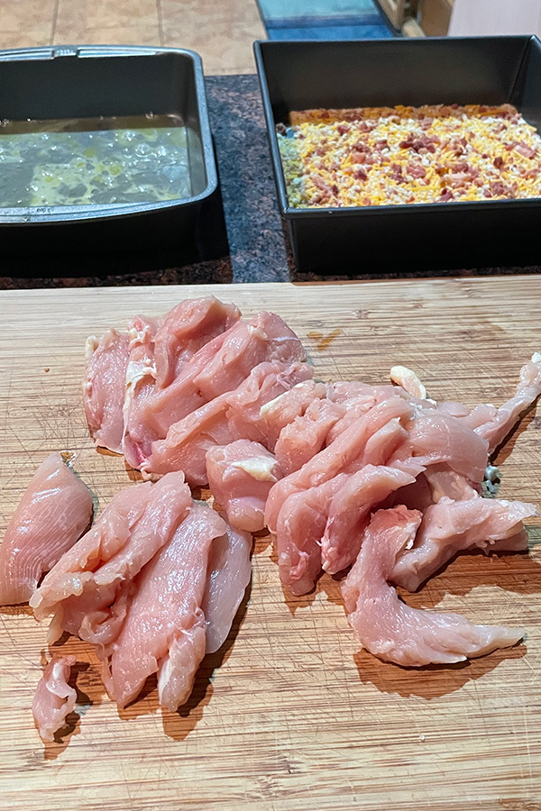 Raw chicken pieces on cutting board, with cheddar bacon bread crumb mixture and whisked egg in cake pans.