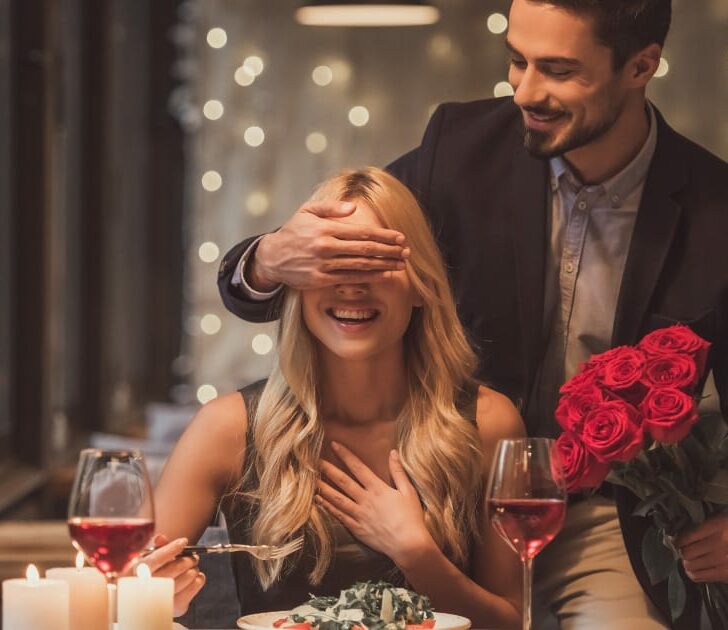 Man stands behind woman who's seated at table set for dining for two.