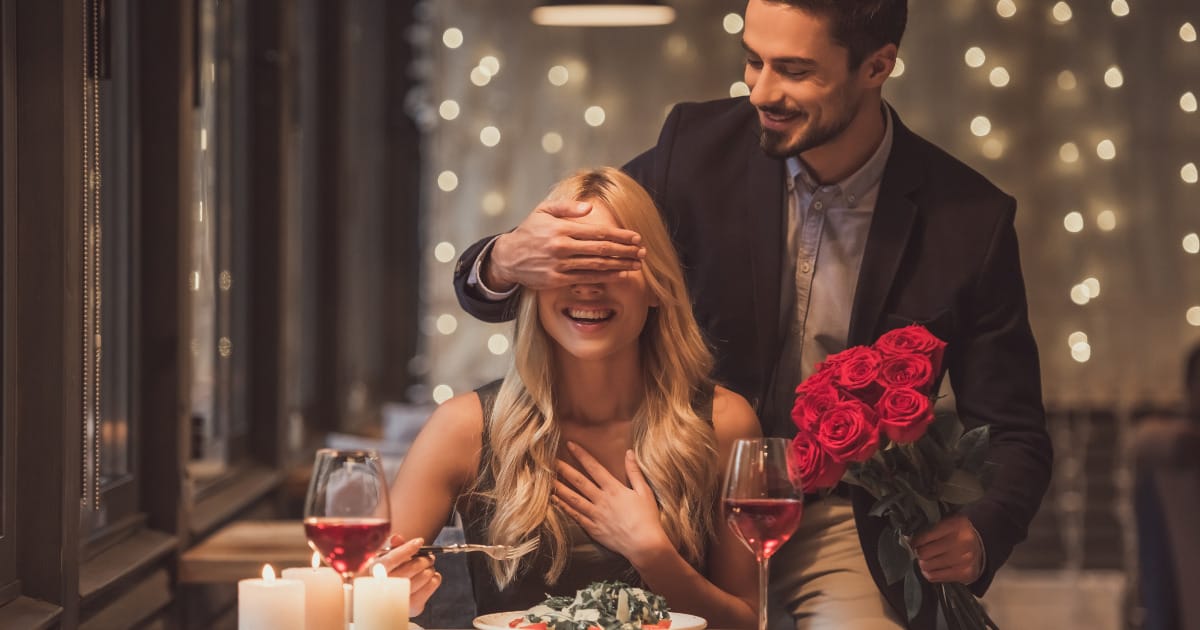 Man stands behind woman who's seated at table set for dining for two.