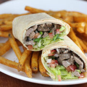 Steak wrap cut in half with lettuce, tomato, and steak filling served with seasoned fries on a white plate