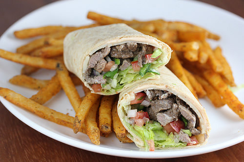 Steak wrap cut in half with lettuce, tomato, and steak filling served with seasoned fries on a white plate