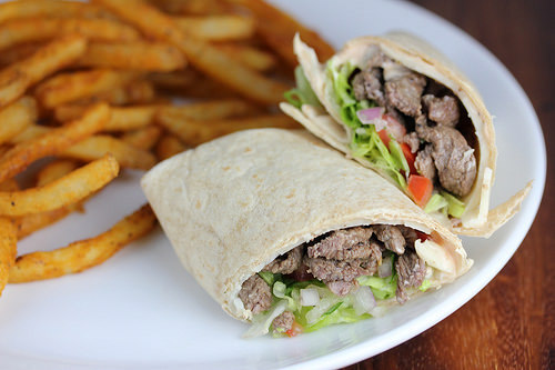 Steak wrap cut in half with lettuce, tomato, and steak filling served with seasoned fries on a white plate