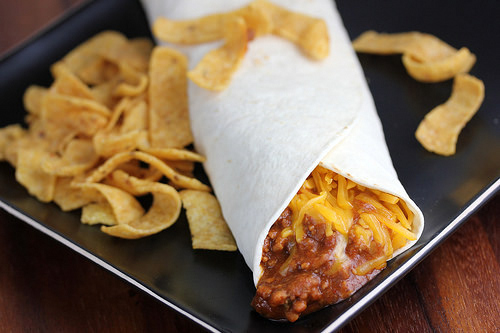 Chili cheese burrito with shredded cheese served with corn chips on a black plate