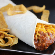 Chili cheese burrito with shredded cheese served with corn chips on a black plate