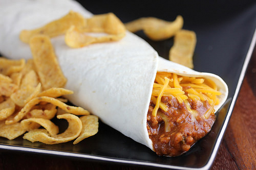 Chili cheese burrito with shredded cheese served with corn chips on a black plate
