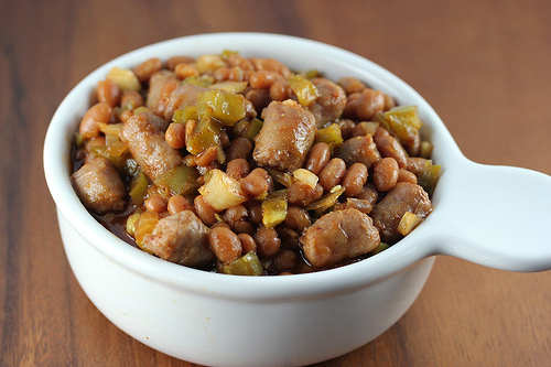 Bowl of spicy baked beans with sausage and diced vegetables on a wooden table