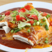 Plate of wet burritos topped with red sauce, melted cheese, lettuce, tomatoes, and green onions