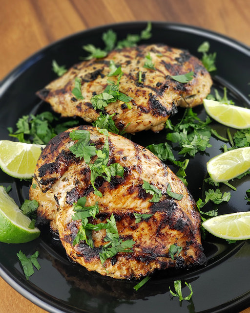 Grilled chicken breasts with cilantro and lime wedges on a black plate